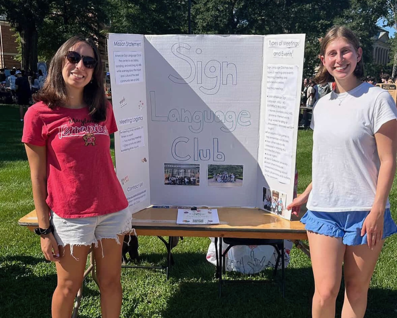 Two students stand outdoors on a grassy campus lawn beside a table displaying a tri-fold poster titled “Sign Language Club,” smiling at the camera during a campus involvement or activities fair.