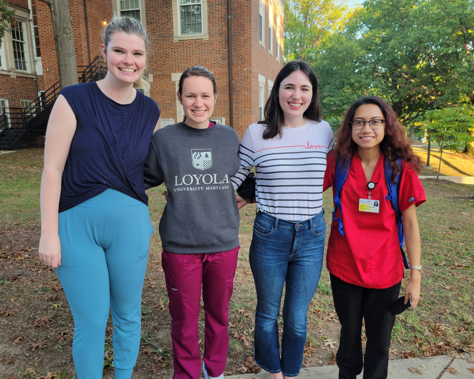 Four students stand together outdoors smiling at the camera, with a brick campus building and trees behind them, wearing a mix of casual clothing and clinical attire.