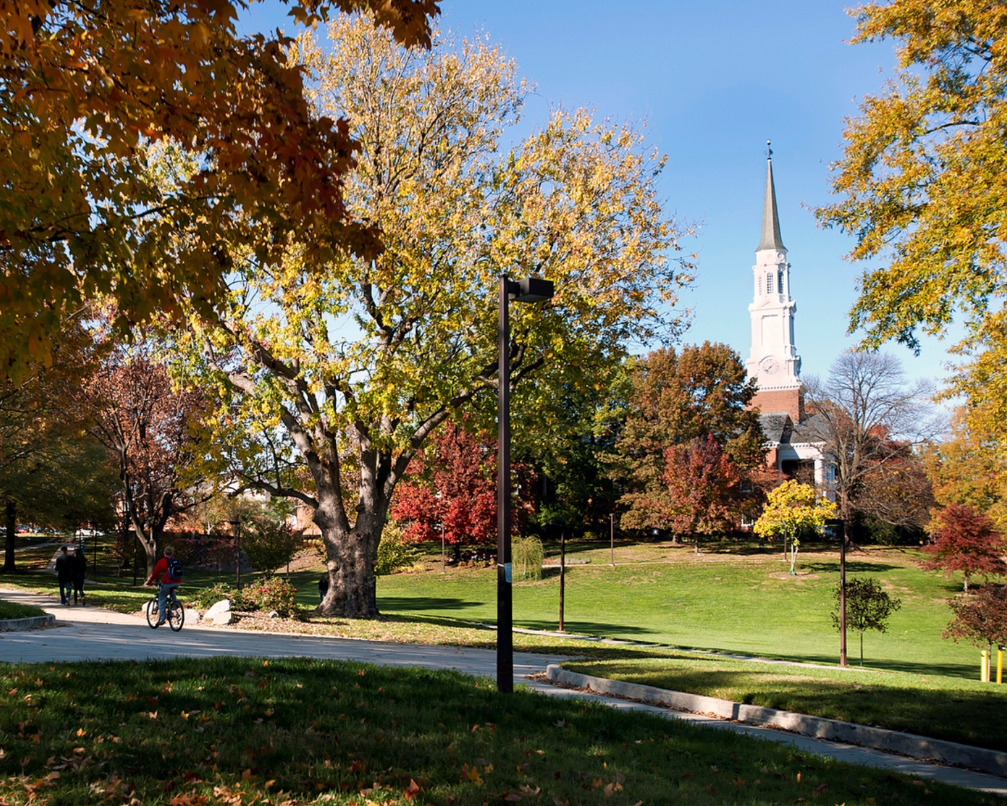 A university campus scene in autumn with trees in fall foliage lining a walking path, a student riding a bicycle, and the Chapel visible above the green lawn in the background.