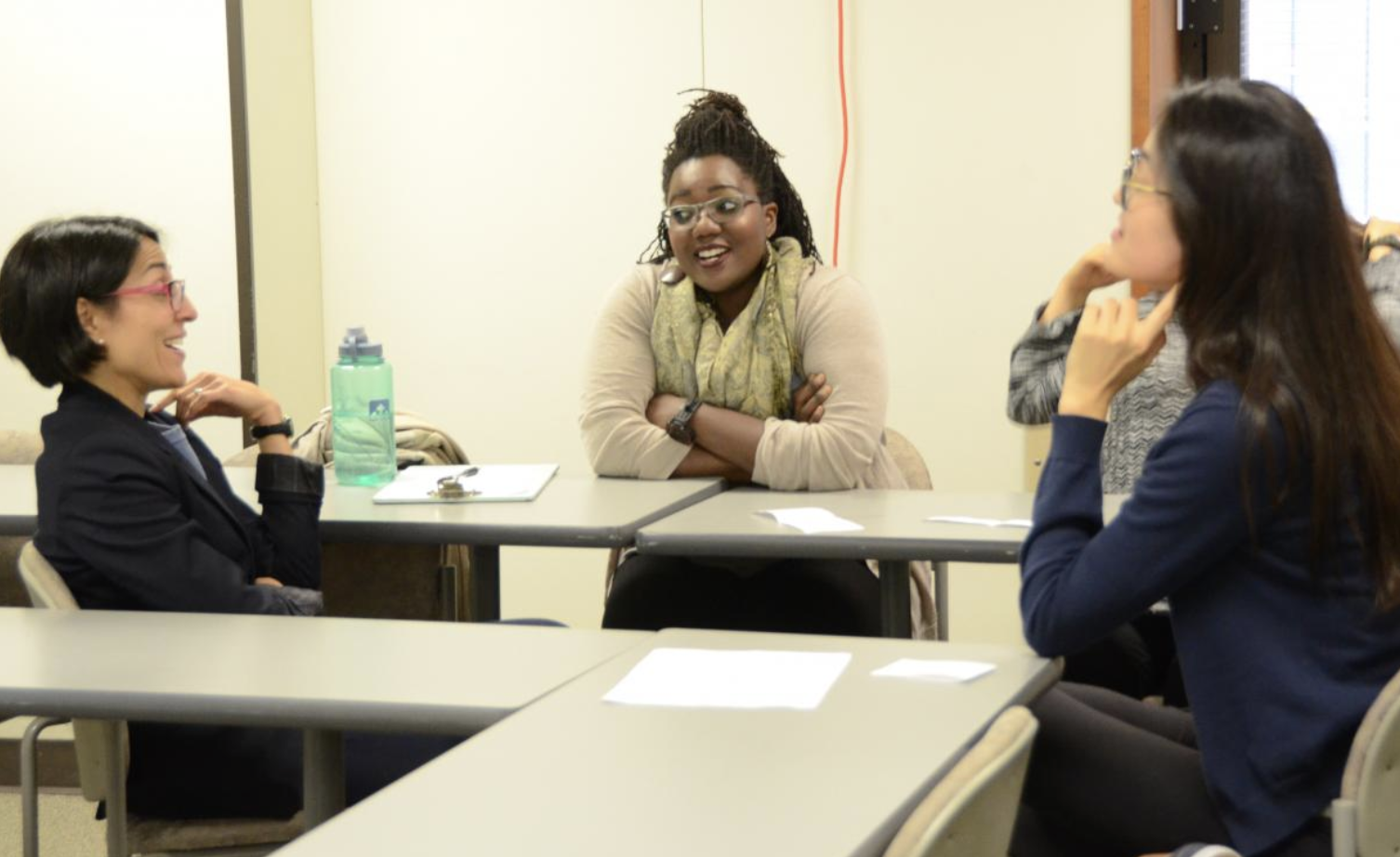 Three students sit around classroom tables in conversation during a small-group discussion; one student speaks while the others listen.
