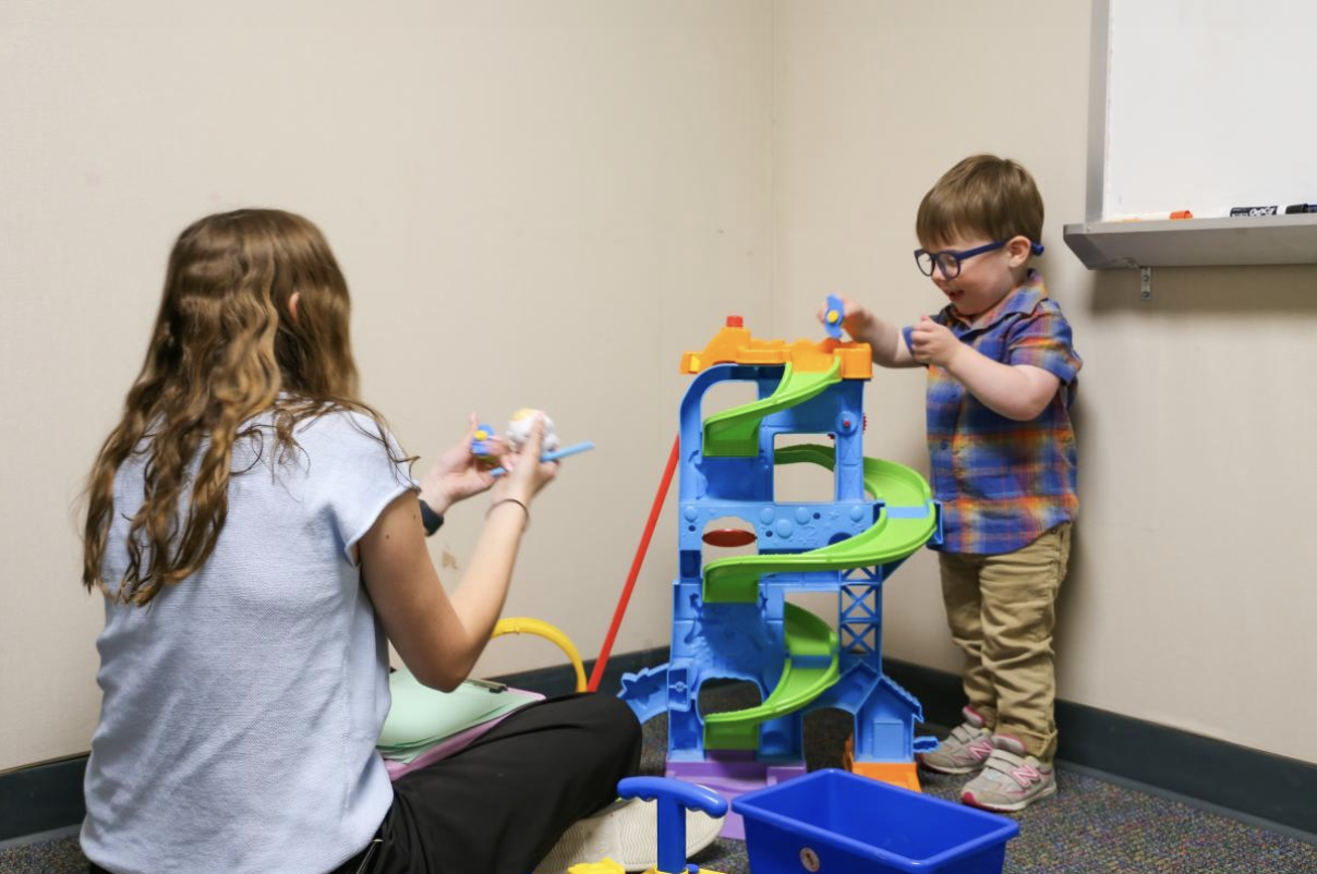 A young child wearing glasses plays with a colorful toy ramp while an adult seated on the floor engages with him in a classroom or therapy setting. A whiteboard is mounted on the wall nearby.