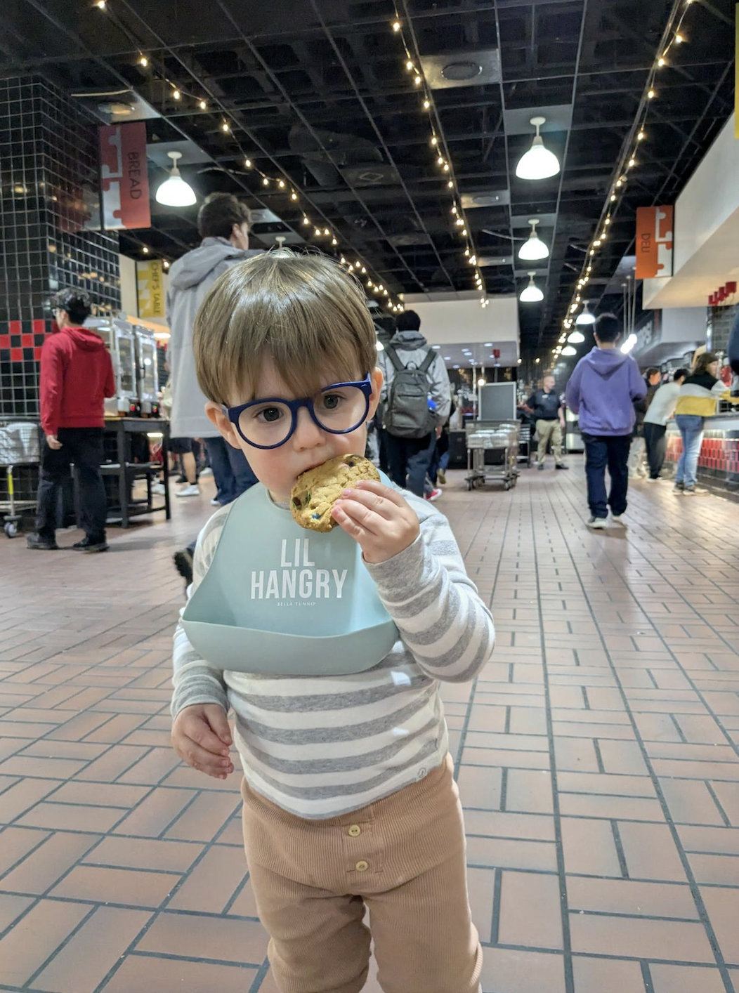 A young child wearing blue glasses and a light blue bib that reads “Lil Hangry” stands in a busy dining hall while eating a cookie. The child is dressed in a gray-and-white striped shirt and tan pants. People walk in the background under string lights and hanging signs, creating a lively indoor food court atmosphere.