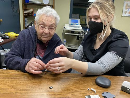 An older woman and a clinician sit together at a wooden table while the clinician helps her handle a small hearing aid. The clinician, wearing scrubs and a black face mask, guides the woman’s hands as they look at the device. Several hearing aid parts and a case sit on the table in front of them. The interaction is part of the JSSA program.