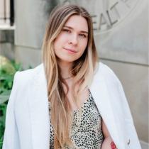 Angelina Bilokon standing outdoors in front of a university building, wearing a patterned top and a white blazer draped over her shoulders. She faces the camera with a slight smile.