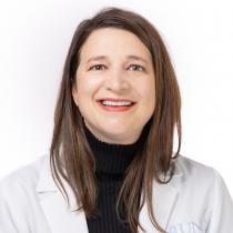 A head-and-shoulders portrait of an adult woman with straight brown hair, wearing a black turtleneck and a white lab coat. She is smiling and facing the camera. The background is plain white.
