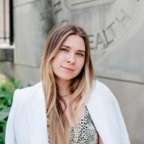 Angelina Bilokon poses outdoors in front of a stone wall engraved with the National Institutes of Health seal. She is wearing a white blazer over a light patterned top and has one arm resting casually at her side. Green plants frame the lower edge of the image.