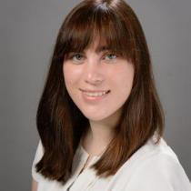 Kelly Marshall, a woman with straight brown hair and bangs, is shown in a studio-style headshot. She is wearing a white blouse and smiling slightly against a plain gray background.