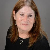 A professional headshot of a woman with shoulder length brown hair wearing a black top, heart shaped necklace, and small earrings. She is smiling softly and posed against a plain gray background.