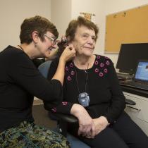 An audiologist examines an older woman’s ear using an otoscope in a clinic room. The patient sits in a desk chair wearing a black top with pink embroidered circles and a small device on a lanyard around her neck. A laptop on the desk beside them displays two graphs representing hearing test results.
