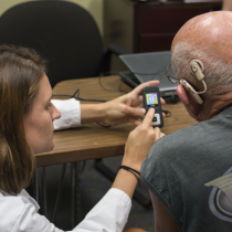 A female audiology clinician sits beside an older man wearing a cochlear implant, showing him how to use a small handheld hearing device. They are seated at a table in a clinical setting with a laptop in the background. The clinician is demonstrating the device’s controls while the patient looks on attentively.