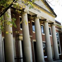A brick academic building with tall white columns and a triangular pediment reading “Samuel J. LeFrak Hall.” Green leaves frame the top left corner of the image, and sunlight filters through the trees, casting shadows on the building’s facade.