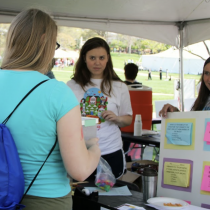 Under a white event tent on a grassy campus lawn, two women stand behind an outreach table speaking with several visitors. A display board on the right shows colorful question cards about the brain and hearing, while a poster behind them appears to promote hearing protection. One visitor in the foreground wears a turquoise shirt and a blue drawstring backpack.