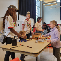 Children and adults gather around a table at a Maryland Day activity indoors, exploring musical instruments with help from student volunteers wearing matching white T-shirts. A young girl in a purple fleece plays an instrument across from a volunteer while other families and children participate in the background.