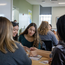 Four students sit around a table in a bright classroom, talking and looking at papers together during a small-group activity.