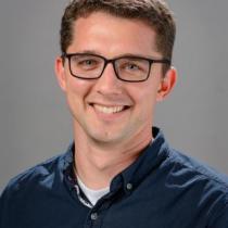 Portrait of a smiling adult man with short brown hair and glasses, wearing a dark blue button-down shirt, photographed against a plain gray background.