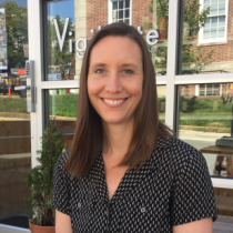 A woman with straight brown hair, wearing a short-sleeve black patterned blouse, sits outside in front of a glass storefront. She is smiling at the camera, and there is a small potted plant beside her.