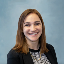 Kelsey Klein smiling in front of a light blue background, wearing a black blazer over a patterned top and a gold chain necklace.