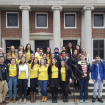 SIGNA students and staff students and staff stand together on the front steps of a brick campus building with tall white columns. Most of the people in the group are smiling and wearing bright yellow T-shirts, while a few are dressed in jackets or sweaters. The group is arranged in several rows and appears to be posing for a group photo outside the entrance.