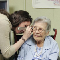 A clinician leans in to examine an older adult’s ear with an otoscope in a clinic room; the older adult sits in a chair and looks ahead.