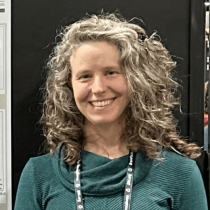 Goldie McQuaid smiling  in front of a black presentation board with scientific charts and graphs. She is wearing a teal sweater and a conference lanyard, appearing to present research at an academic event.