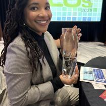 Dr. Courtney Overton in a gray checkered suit sits at a round table, smiling while holding a clear crystal award. She has long locs and is seated in front of a large projected screen that reads “Maryland’s Most Admired CEOs 2025.” The room appears to be a formal event setting with programs and papers on the table.