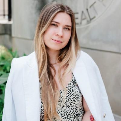 Angelina Bilokon standing outdoors in front of a university building, wearing a patterned top and a white blazer draped over her shoulders. She faces the camera with a slight smile.