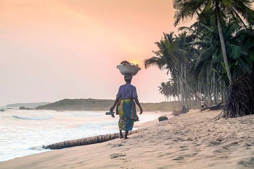 A person walks barefoot along a sandy beach at sunset, carrying a large bowl balanced on their head and holding a pair of sandals. Palm trees line the shore, and gentle waves roll in under a pink and orange sky.
