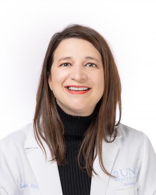 A head-and-shoulders portrait of an adult woman with straight brown hair, wearing a black turtleneck and a white lab coat. She is smiling and facing the camera. The background is plain white.