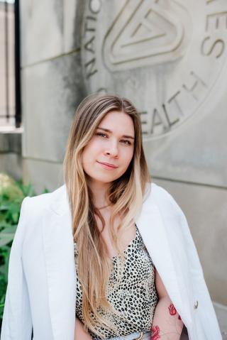 A woman with blonde hair wears a lab coat and looks at the camera. She is outside. 
