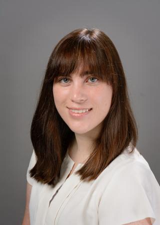 Kelly Marshall, a woman with straight brown hair and bangs, is shown in a studio-style headshot. She is wearing a white blouse and smiling slightly against a plain gray background.