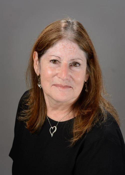 A professional headshot of a woman with shoulder length brown hair wearing a black top, heart shaped necklace, and small earrings. She is smiling softly and posed against a plain gray background.