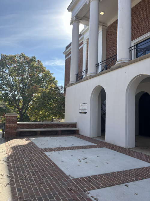 A brick and white-stucco university building with tall columns and arched entrances on a sunny day. A tree with early fall foliage stands to the left, and scattered leaves lie on the brick walkway in front of the building. A sign next to the entrance identifies the location as LeFrak Hall at the University of Maryland.
