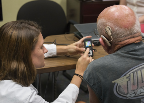 A female audiology clinician sits beside an older man wearing a cochlear implant, showing him how to use a small handheld hearing device. They are seated at a table in a clinical setting with a laptop in the background. The clinician is demonstrating the device’s controls while the patient looks on attentively.