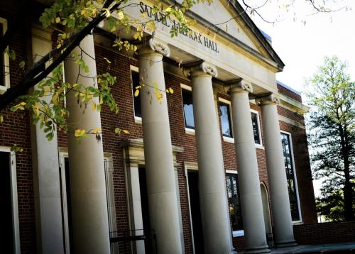 A brick academic building with tall white columns and a triangular pediment reading “Samuel J. LeFrak Hall.” Green leaves frame the top left corner of the image, and sunlight filters through the trees, casting shadows on the building’s facade.