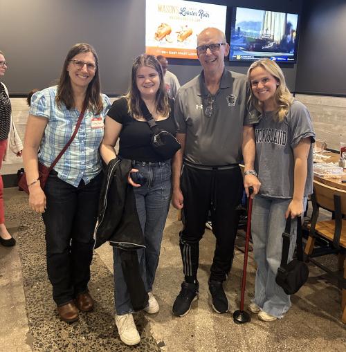 Four people stand indoors posing for a photo. An older man using a cane is standing between two younger women. A woman in glasses stands on the far left. They are all smiling. The background shows a restaurant-style setting with tables and digital menu screens.