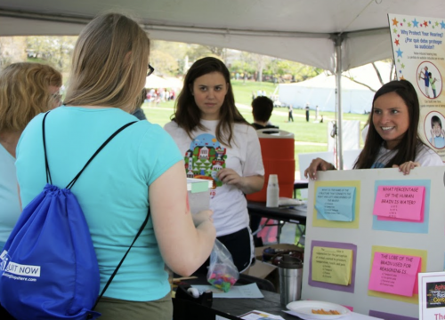 Under a white event tent on a grassy campus lawn, two women stand behind an outreach table speaking with several visitors. A display board on the right shows colorful question cards about the brain and hearing, while a poster behind them appears to promote hearing protection. One visitor in the foreground wears a turquoise shirt and a blue drawstring backpack.