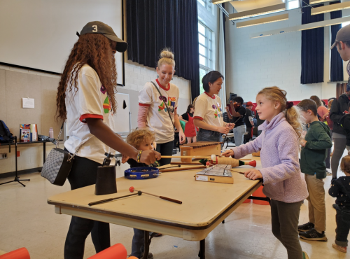 Children and adults gather around a table at a Maryland Day activity indoors, exploring musical instruments with help from student volunteers wearing matching white T-shirts. A young girl in a purple fleece plays an instrument across from a volunteer while other families and children participate in the background.