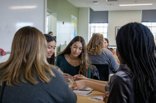 Four students sit around a table in a bright classroom, talking and looking at papers together during a small-group activity.