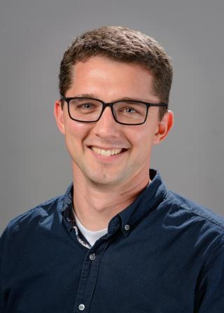 Portrait of a smiling adult man with short brown hair and glasses, wearing a dark blue button-down shirt, photographed against a plain gray background.