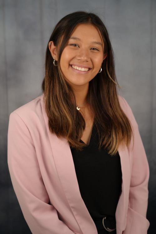 Headshot of a smiling person with long brown hair wearing a light pink blazer over a black top, standing in front of a gray backdrop.