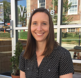A woman with straight brown hair, wearing a short-sleeve black patterned blouse, sits outside in front of a glass storefront. She is smiling at the camera, and there is a small potted plant beside her.