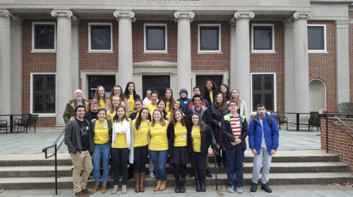SIGNA students and staff students and staff stand together on the front steps of a brick campus building with tall white columns. Most of the people in the group are smiling and wearing bright yellow T-shirts, while a few are dressed in jackets or sweaters. The group is arranged in several rows and appears to be posing for a group photo outside the entrance.