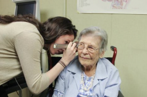 A clinician leans in to examine an older adult’s ear with an otoscope in a clinic room; the older adult sits in a chair and looks ahead.