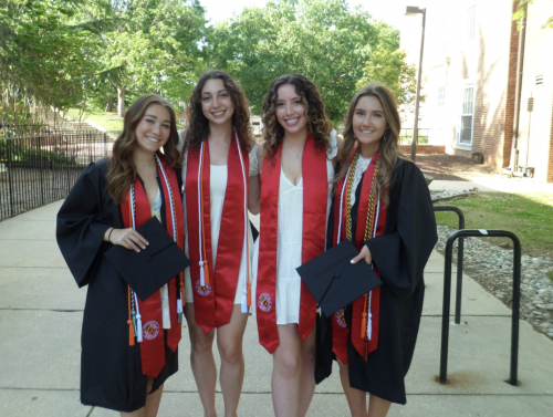 Four smiling graduates stand together outdoors on a campus walkway, wearing black graduation gowns and red stoles while holding their caps. Trees and campus buildings are visible in the background on a sunny day.