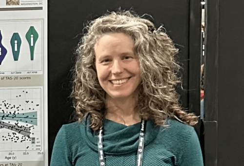 Goldie McQuaid smiling in front of a black presentation board with scientific charts and graphs. She is wearing a teal sweater and a conference lanyard, appearing to present research at an academic event.