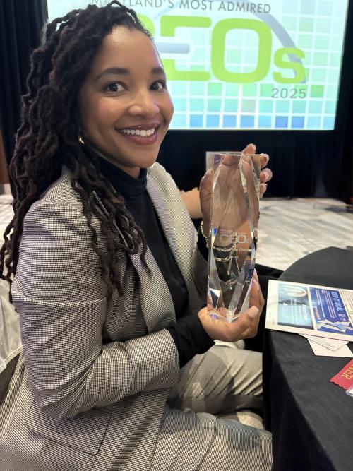 Dr. Courtney Overton in a gray checkered suit sits at a round table, smiling while holding a clear crystal award. She has long locs and is seated in front of a large projected screen that reads “Maryland’s Most Admired CEOs 2025.” The room appears to be a formal event setting with programs and papers on the table.