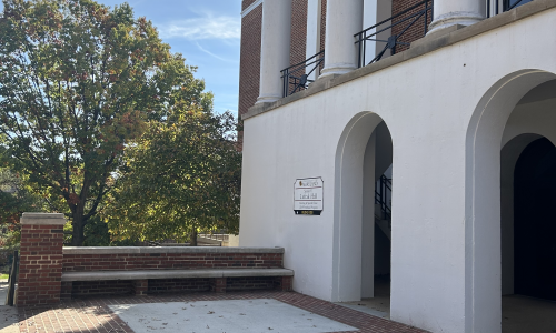 A brick and white-stucco university building with tall columns and arched entrances on a sunny day. A tree with early fall foliage stands to the left, and scattered leaves lie on the brick walkway in front of the building. A sign next to the entrance identifies the location as LeFrak Hall at the University of Maryland.