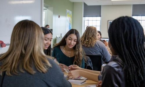 Several students sit around a table in a bright classroom, talking and looking at papers together during a small group activity. A whiteboard is on the left wall, and more students are seated at tables in the background near large windows.