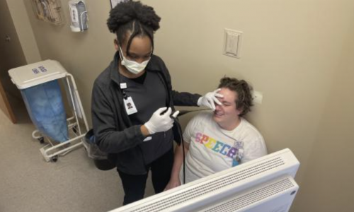A student clinician wearing gloves and a face mask performs a FEES examination on a seated patient in a clinic room while the patient smiles. A computer monitor is visible in the foreground, and medical supplies are nearby.