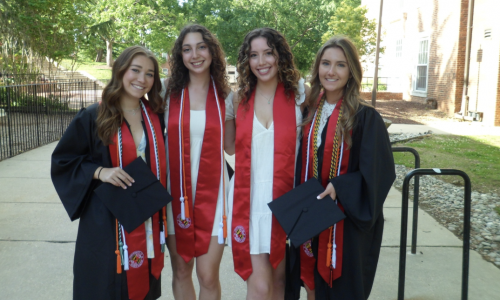Four smiling graduates stand together outdoors on a campus walkway, wearing black graduation gowns and red stoles while holding their caps. Trees and campus buildings are visible in the background on a sunny day.
